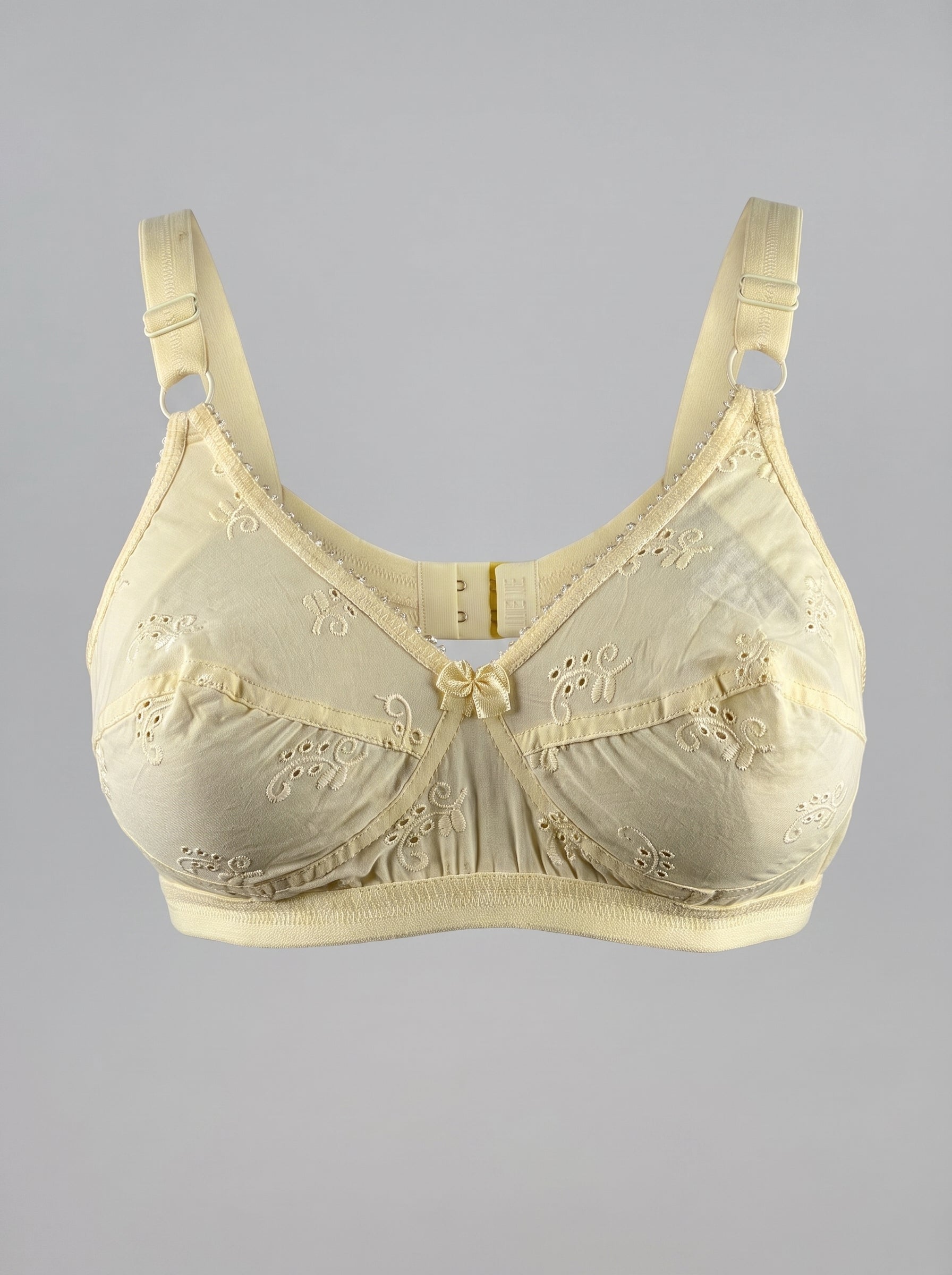 A pale yellow underwire bra with intricate lace and eyelet embroidery, featuring adjustable shoulder straps and a center bow, shown against a solid gray background. #color_cream