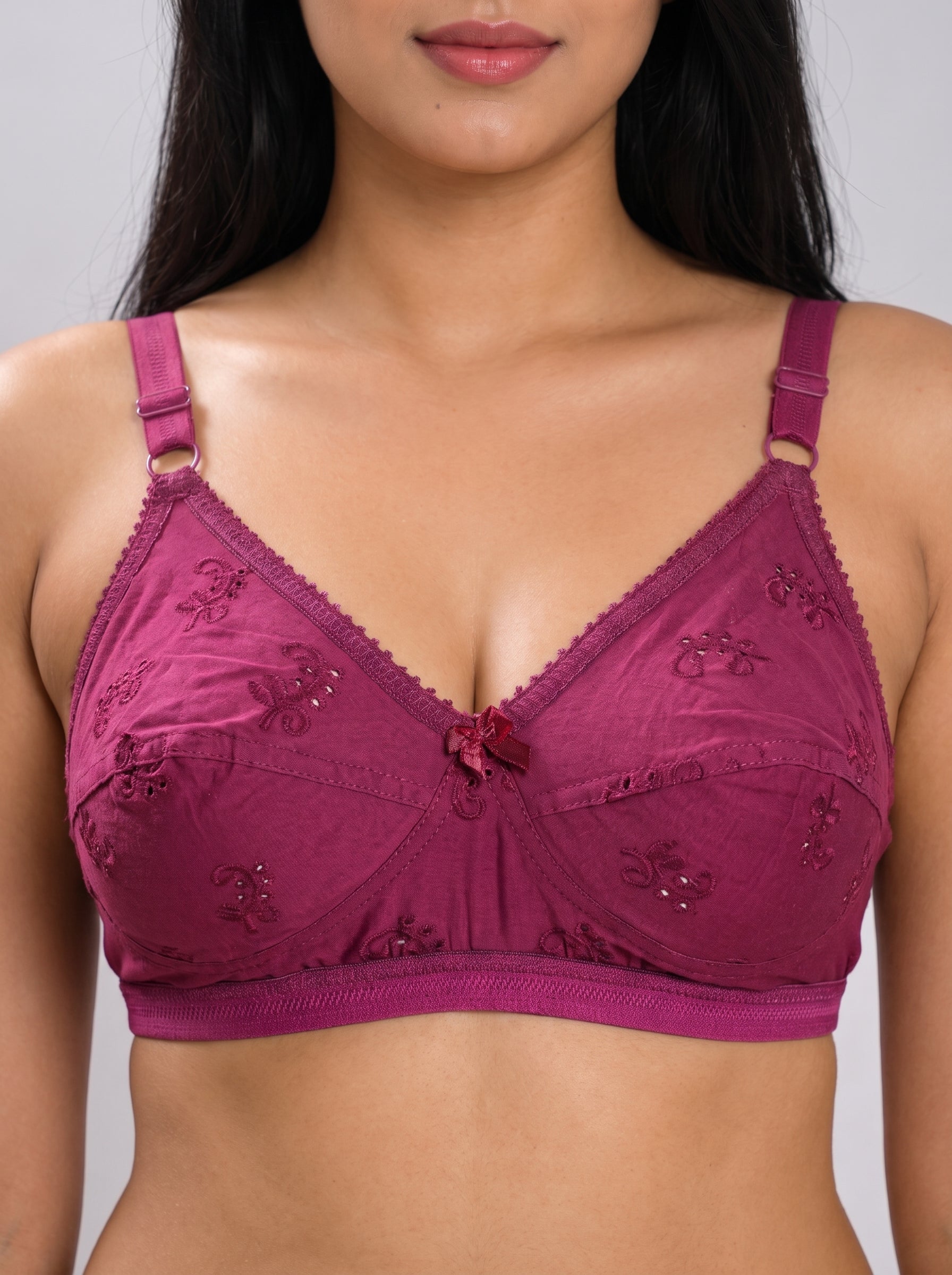 A close-up of a woman wearing a magenta lace bra with embroidered floral details and a small bow at the center, set against a plain gray background. #color_maroon