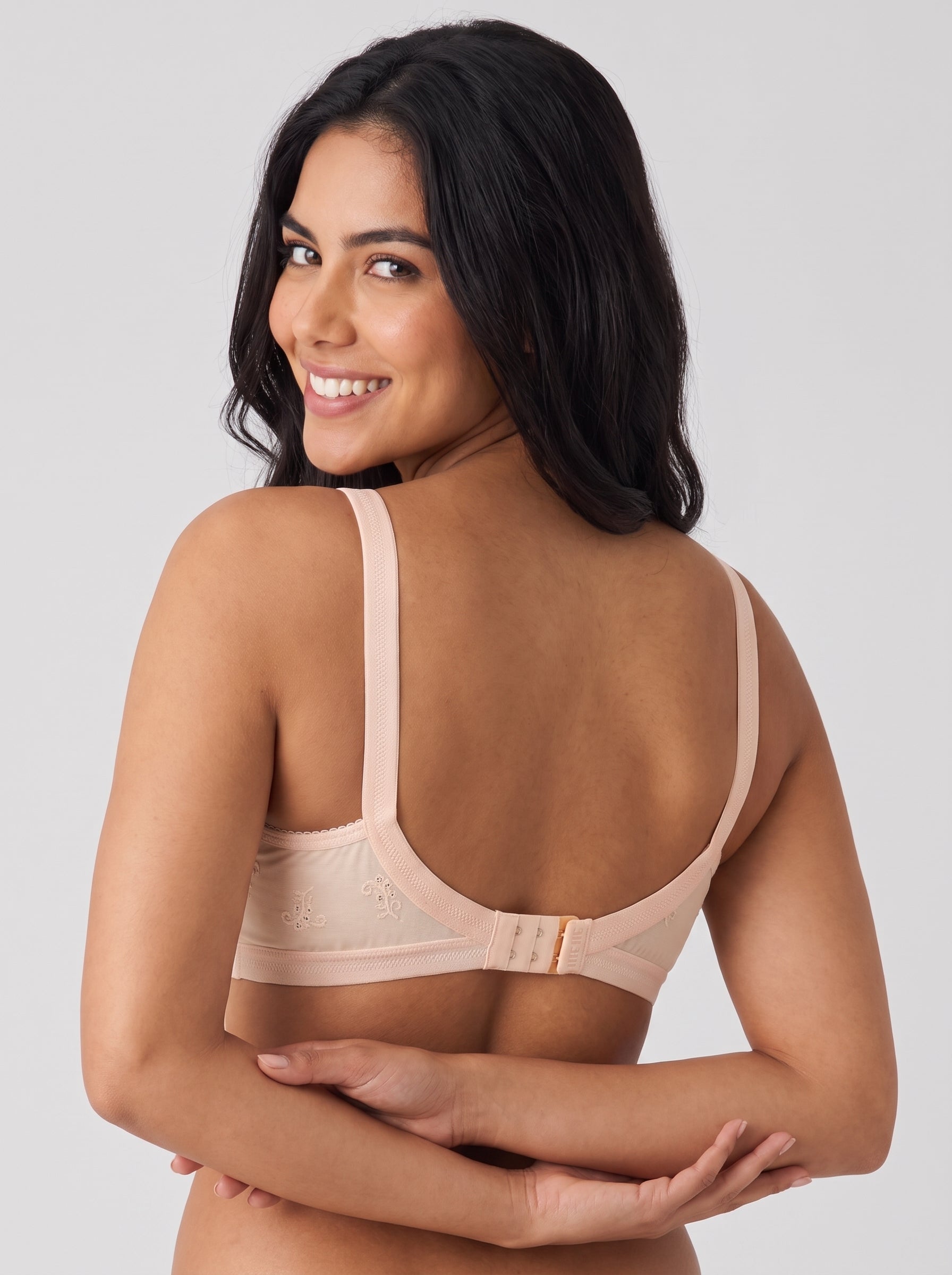 A woman with long dark hair is shown from the back wearing a light beige underwire bra against a solid light gray studio background. #color_peach