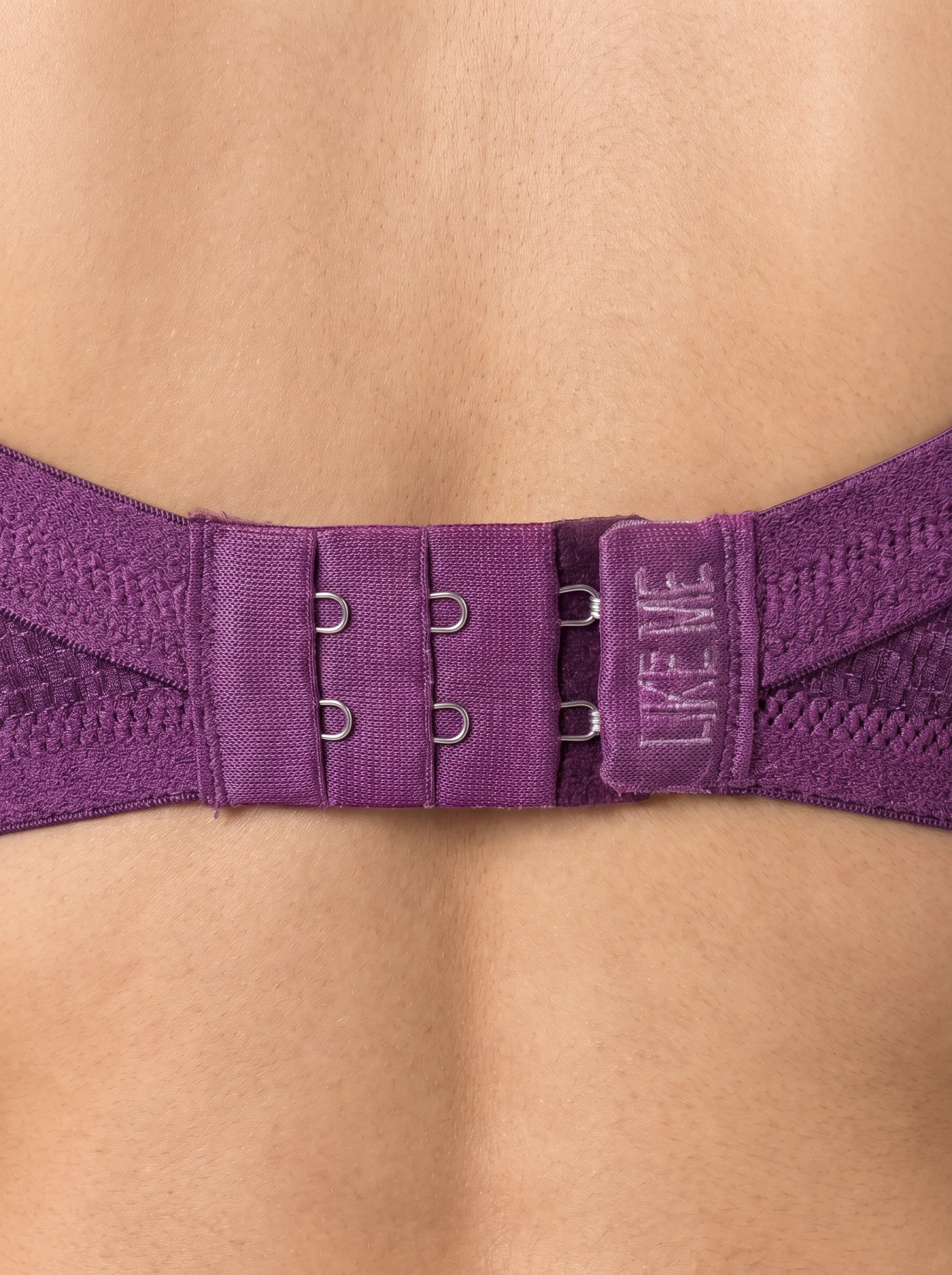 Close-up of a purple bra back strap with lace trim, metal adjusters, and a hook-and-loop closure tab that reads 'LIKE ME' in white text, worn on a model's back. #color_wine
