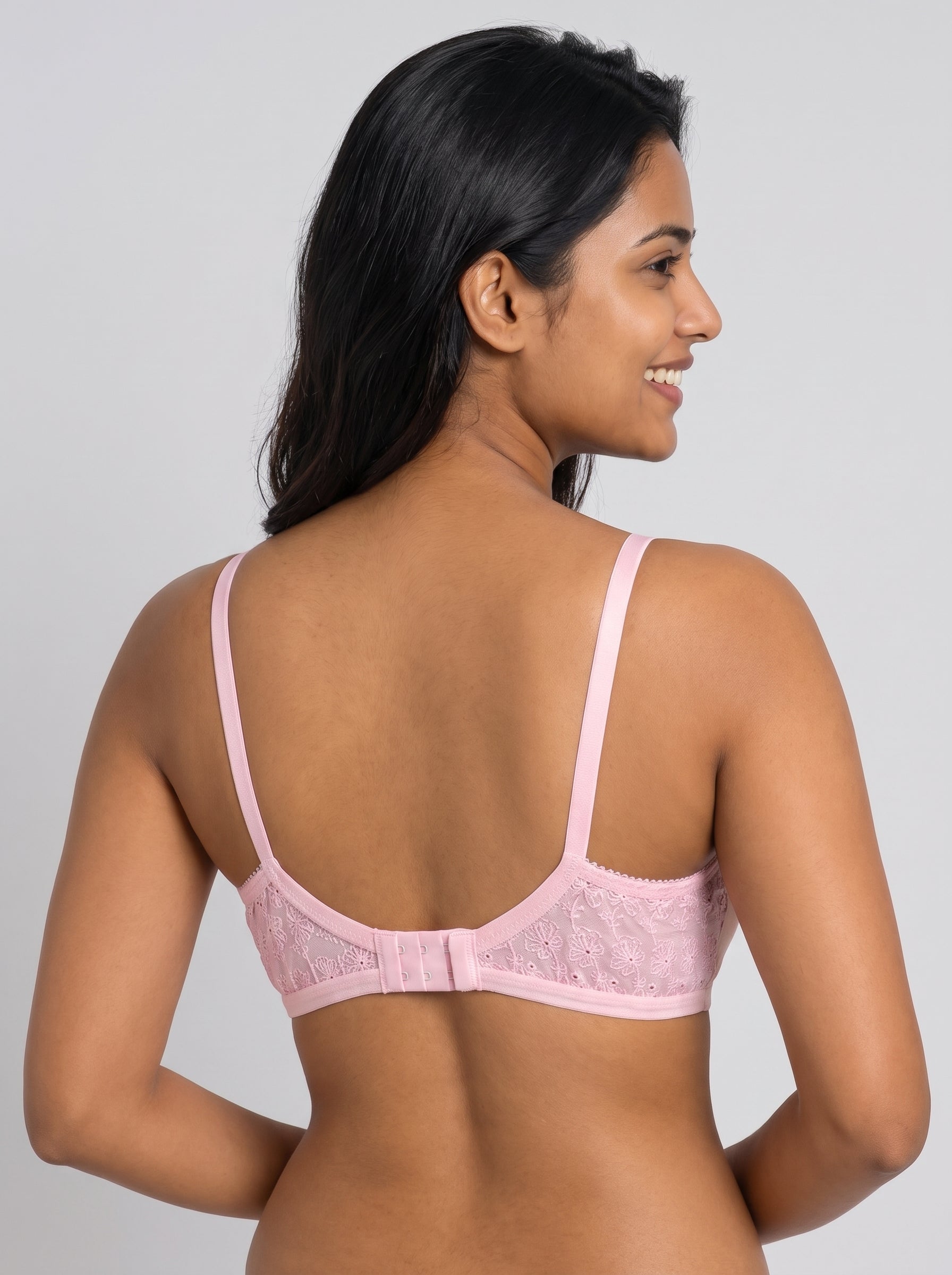 A woman shown from the back wearing a light pink lace bra with thin straps and a center back strap, against a plain light gray studio background. #color_pink