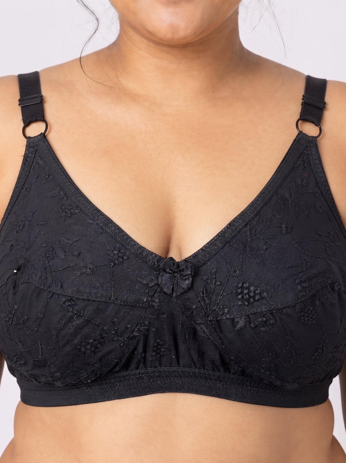 Close-up of Like-Me Sonu Bra in black on plus-size model, showing floral lace cups, center bow and supportive underband. #color_black