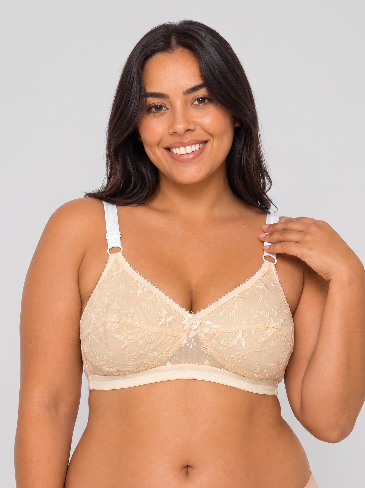 A smiling woman with dark hair models a beige lace underwire bra with white adjustable shoulder straps against a light gray studio background. #color_skin