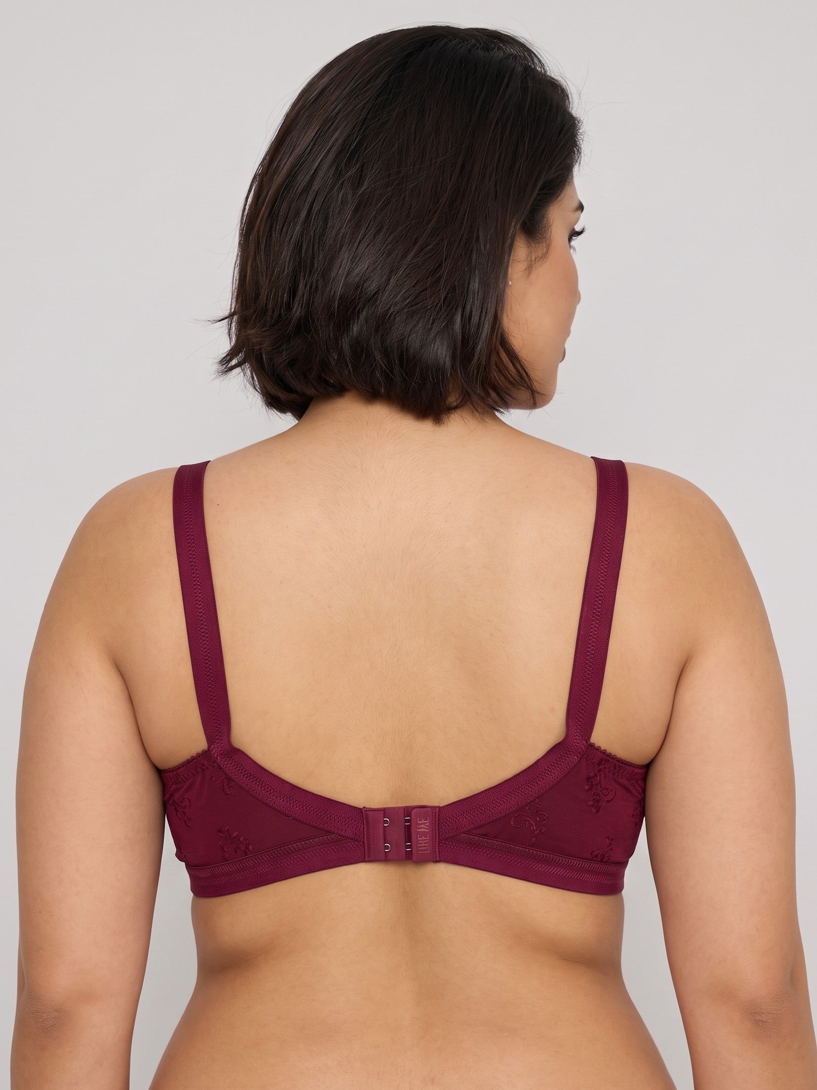 A woman shown from the back wearing a burgundy lace bra with wide shoulder straps and a center back strap closure, against a plain light gray studio background. #color_maroon