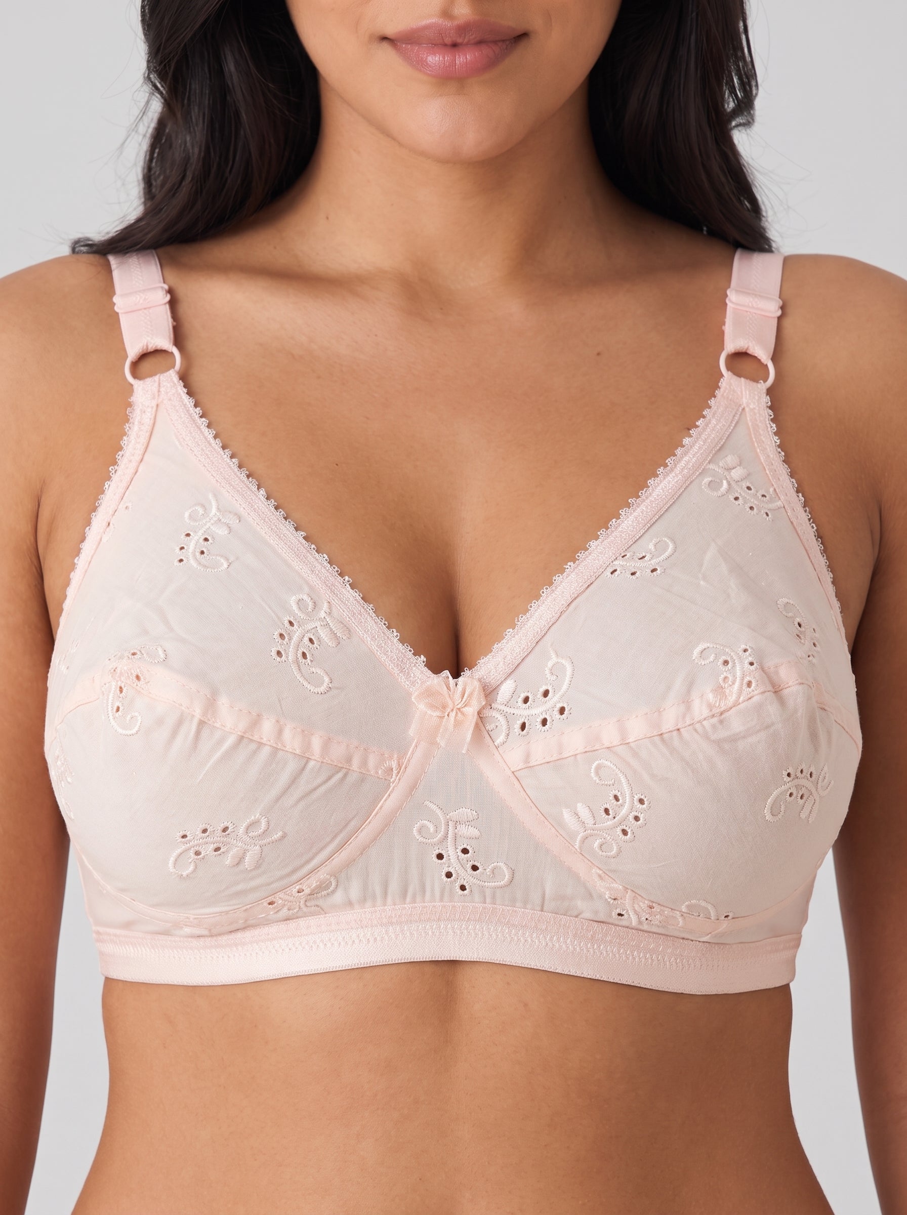 A close-up of a model wearing a light pink underwire bra with eyelet lace detailing and adjustable shoulder straps. #color_peach