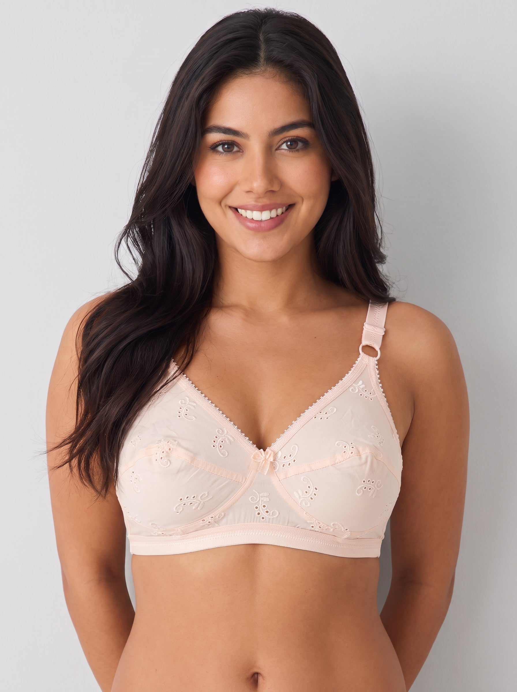 A smiling woman with long dark hair wearing a pale blush pink underwire bra with floral eyelet lace and adjustable shoulder straps, photographed against a light gray studio background. #color_peach