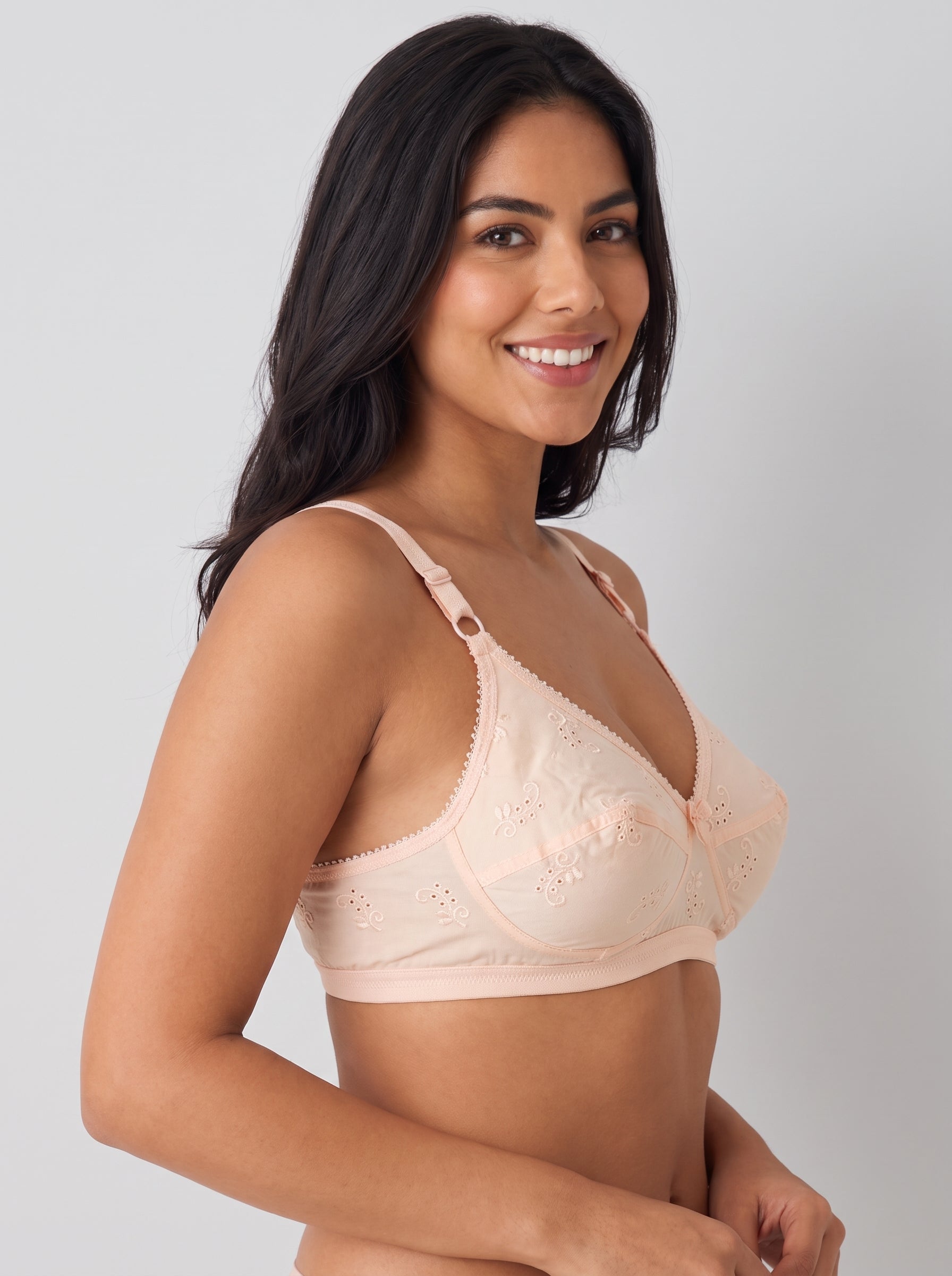A smiling woman with long dark hair models a light peach-beige lace bra against a plain light gray studio background. #color_peach