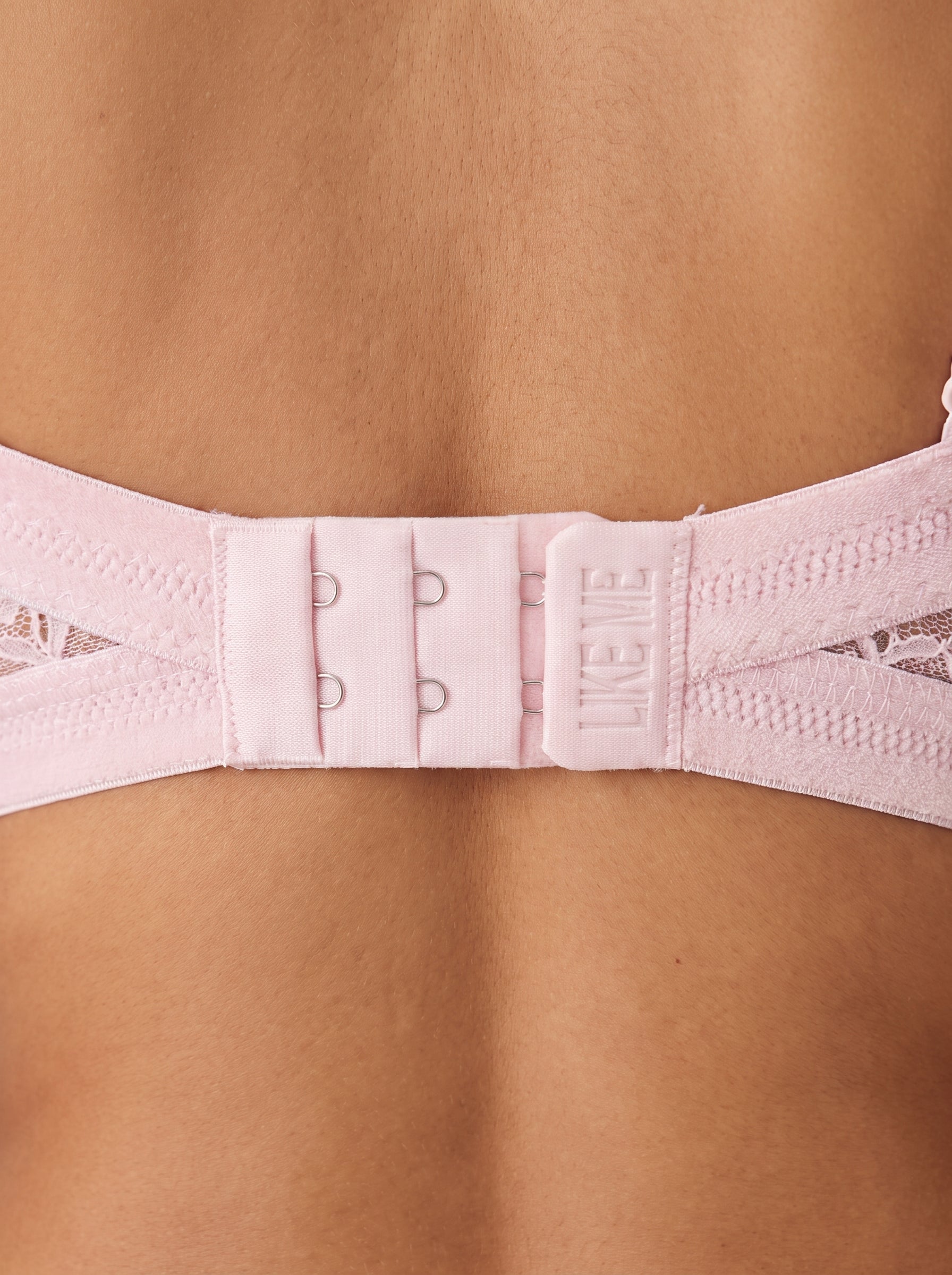 Close-up of a light pink bra strap adjustment panel worn on a model's back, showing the hook-and-eye closure and the brand name 'LIKE ME' printed on the central tab. #color_pink
