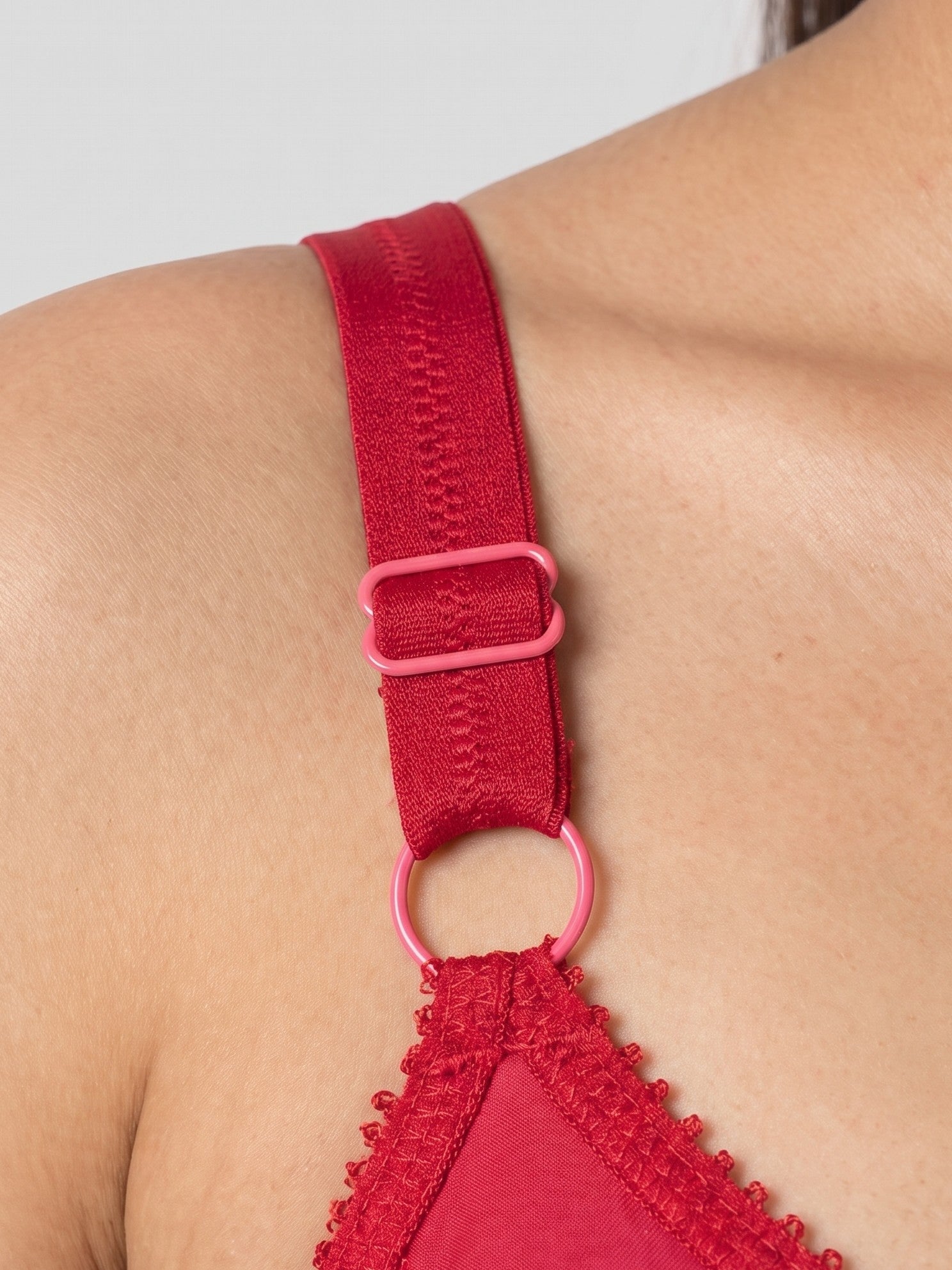 Close-up of a red bra strap with a pink plastic adjuster and ring detail, worn on a model's shoulder. #color_red