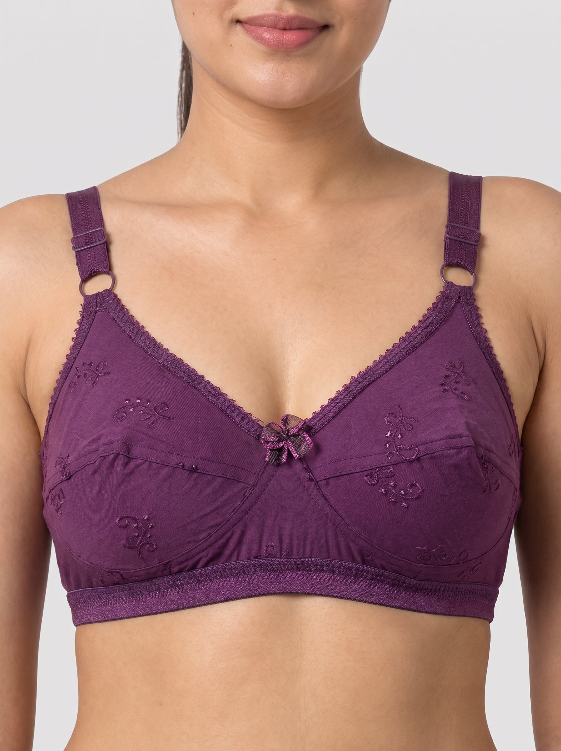 A woman wearing a purple underwire bra with lace detailing and a small bow at the center front, shown against a light gray studio background. #color_wine