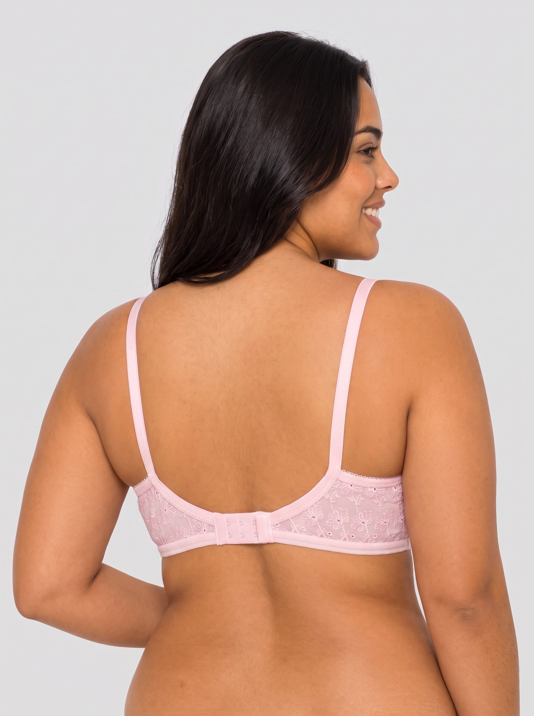 A woman shown from the back wearing a light pink underwire bra with delicate lace detailing on the cups and a wide band across the lower back, set against a solid light gray background. #color_pink