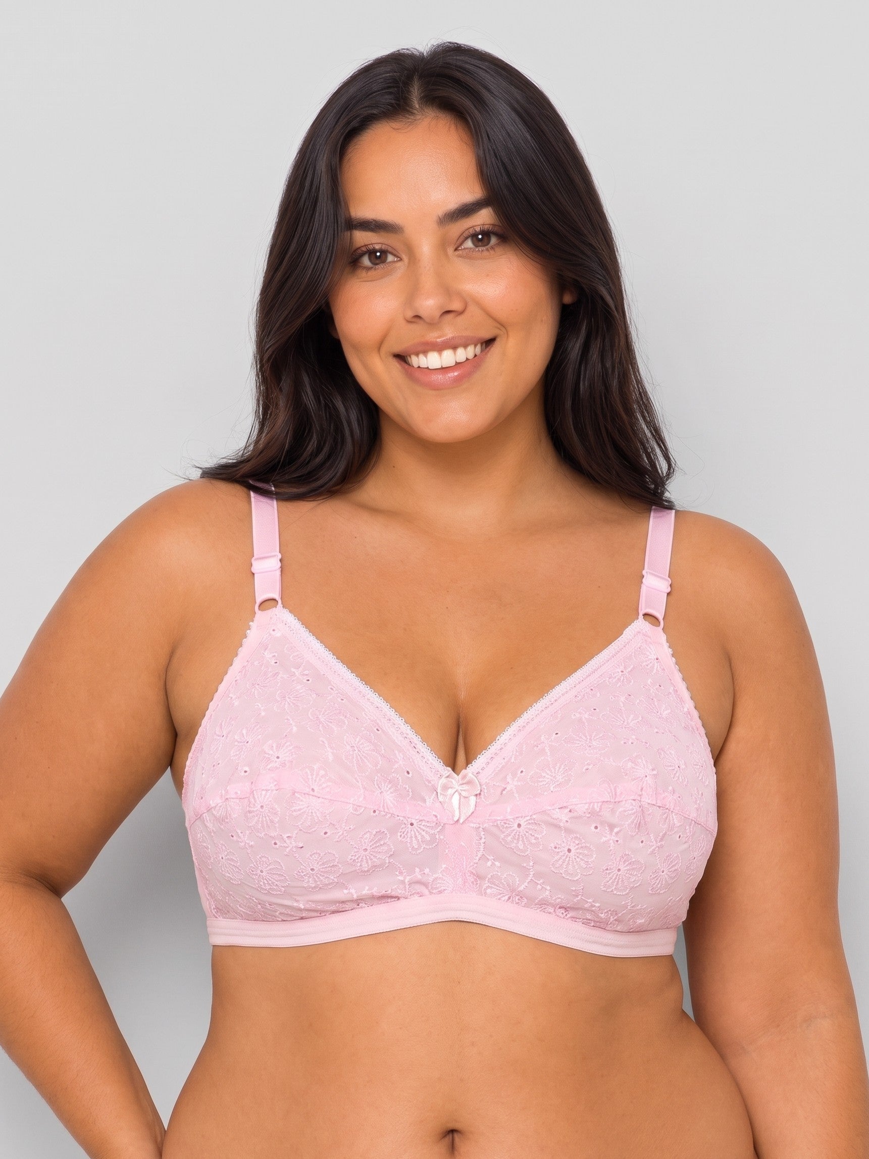 A smiling woman with dark hair wearing a light pink lace bra, shown from the chest up against a plain light gray studio background. #color_pink